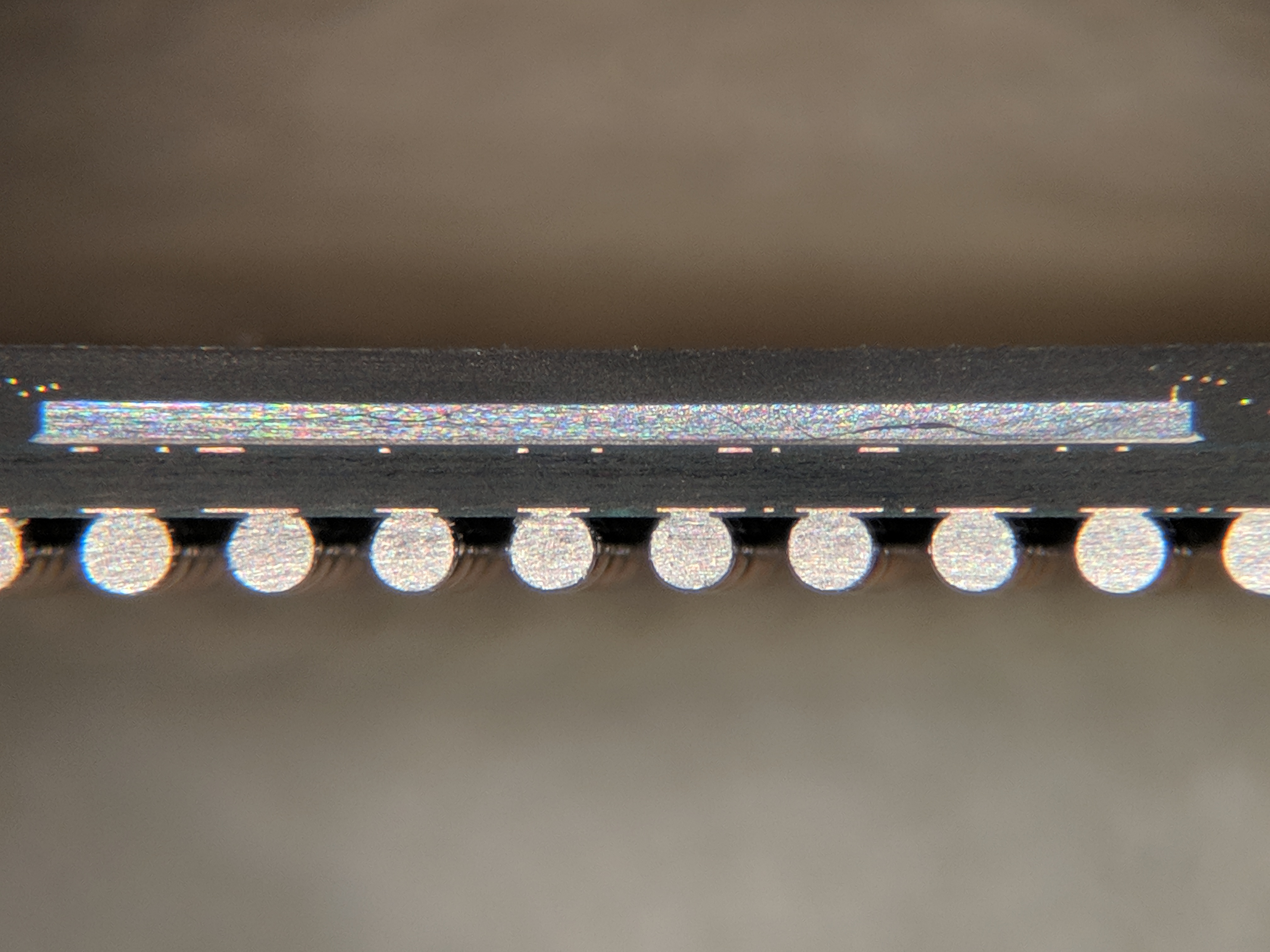 Macro photograph showing a cross-section of a BGA chip package mounted on a PCB. The chip silicon die sits inside a black plastic package on top, the package substrate forms a thin layer in the middle, and a row of round solder balls connects the package to the PCB pads at the bottom. Each solder ball is the size of a fine grain of sand and forms one electrical connection between the chip and the board. To remove the chip, an examiner must liquefy or mechanically separate every one of these solder balls without damaging the silicon die above.